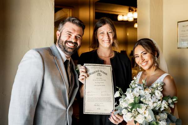 Minister Allison Donohue, an officiant with Texas Wedding Ministers, with a couple at The Bell Tower on 34th wedding venue. Minister Allison Donohue, with Texas Wedding Ministers, with a couple smiling and holding their wedding license at The Bell Tower on 34th wedding venue.